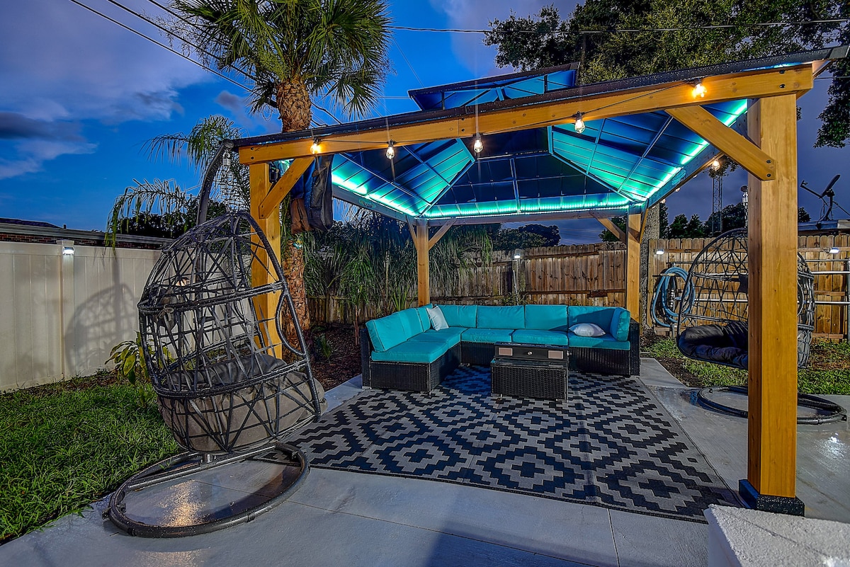 A shaded outdoor gazebo is highlighted by a geometric patterned rug and sectional seating in a calming blue hue. Two hanging egg chairs are positioned for relaxation, while warm lights illuminate the space under a clear, sloped roof, surrounded by tropical plants.