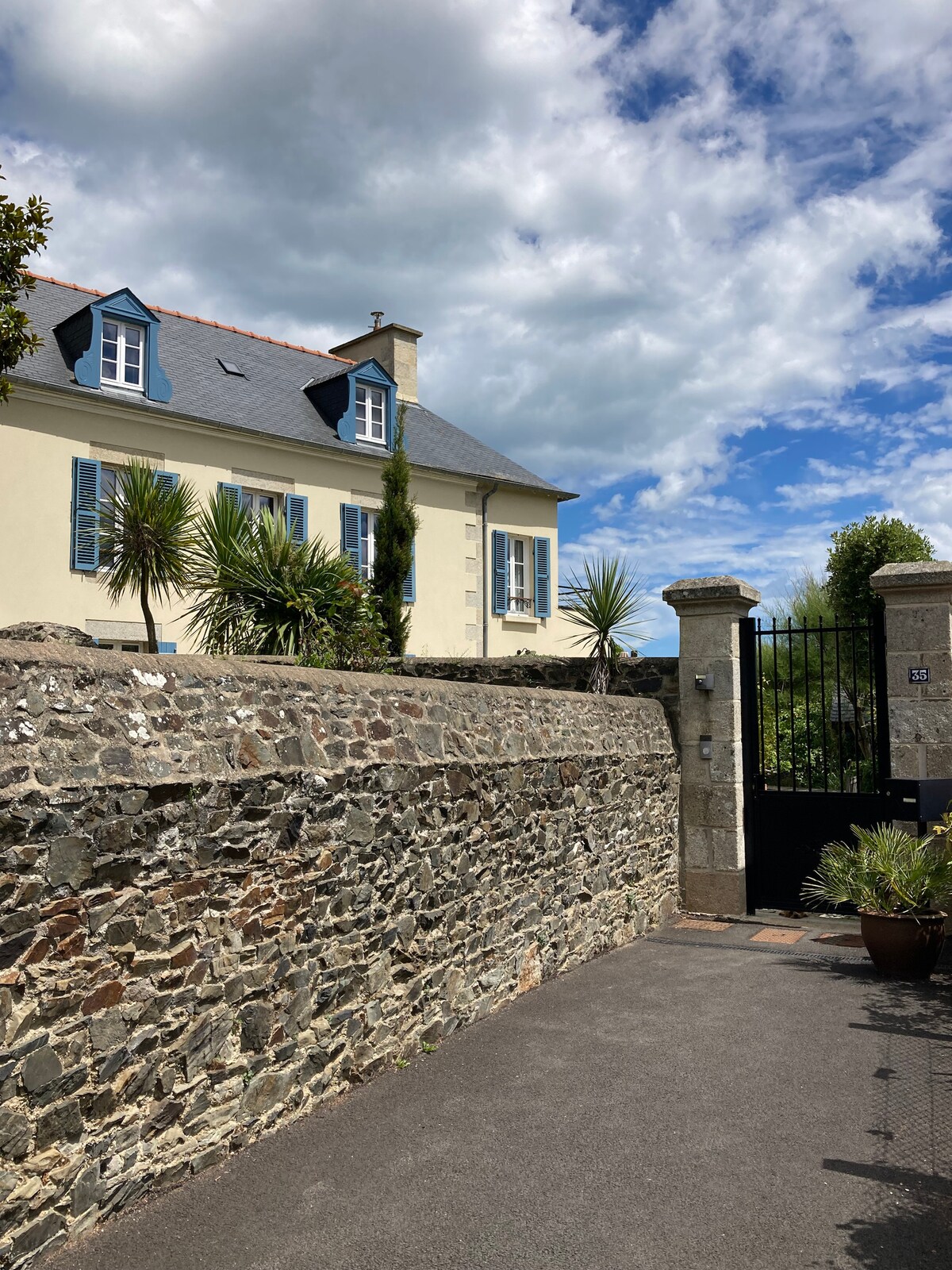 The exterior of a charming two-story house is visible, framed by a stone wall. Lush greenery and palm-like plants add to the welcoming environment. A blue sky with scattered clouds provides a bright backdrop, while a gated entrance is situated to the right, leading into the property.