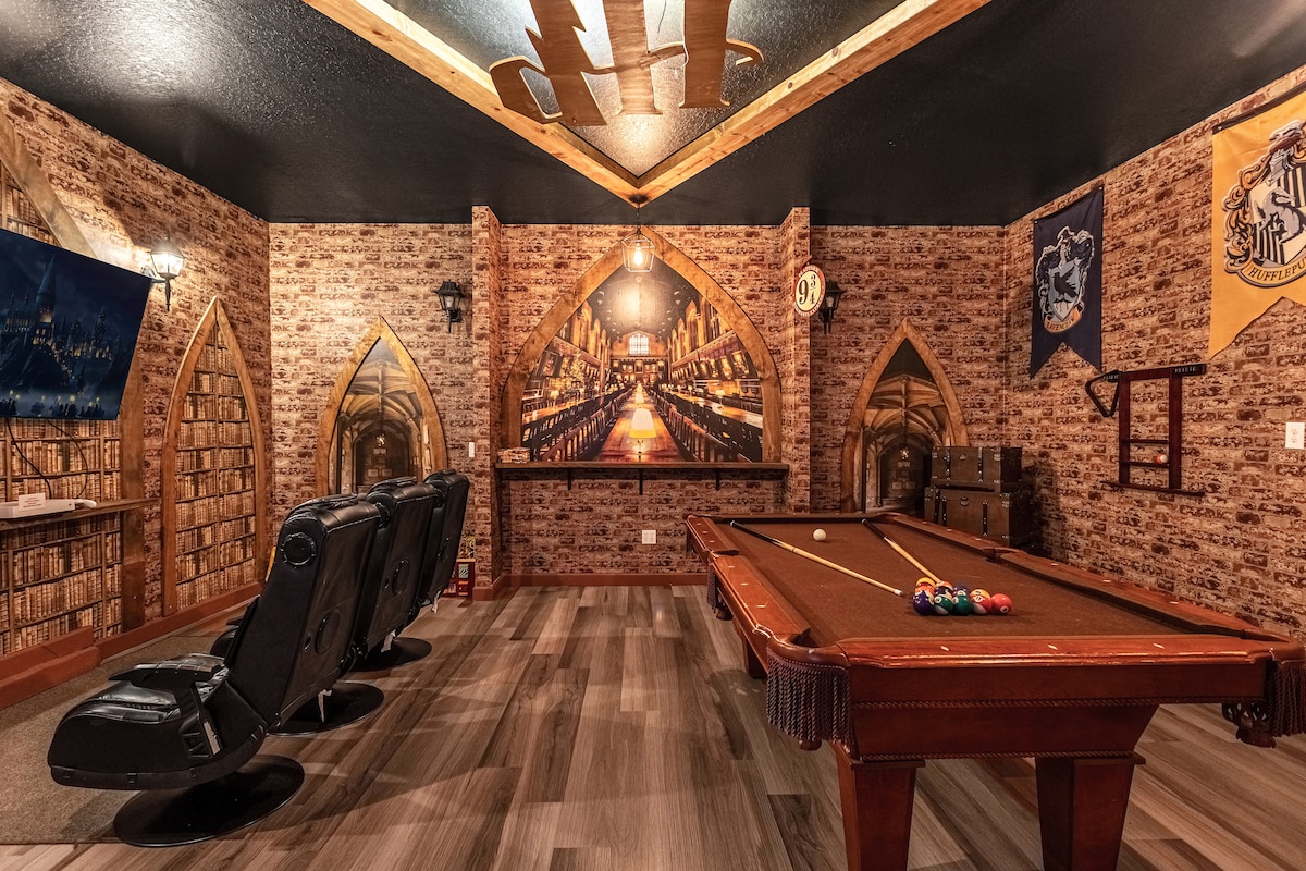 A themed game room features a billiard table with a complete set of pool balls, flanked by leather recliners for seating. The walls are adorned with brick design and Hogwarts-themed banners, creating a cozy atmosphere. Soft lighting enhances the immersive experience.