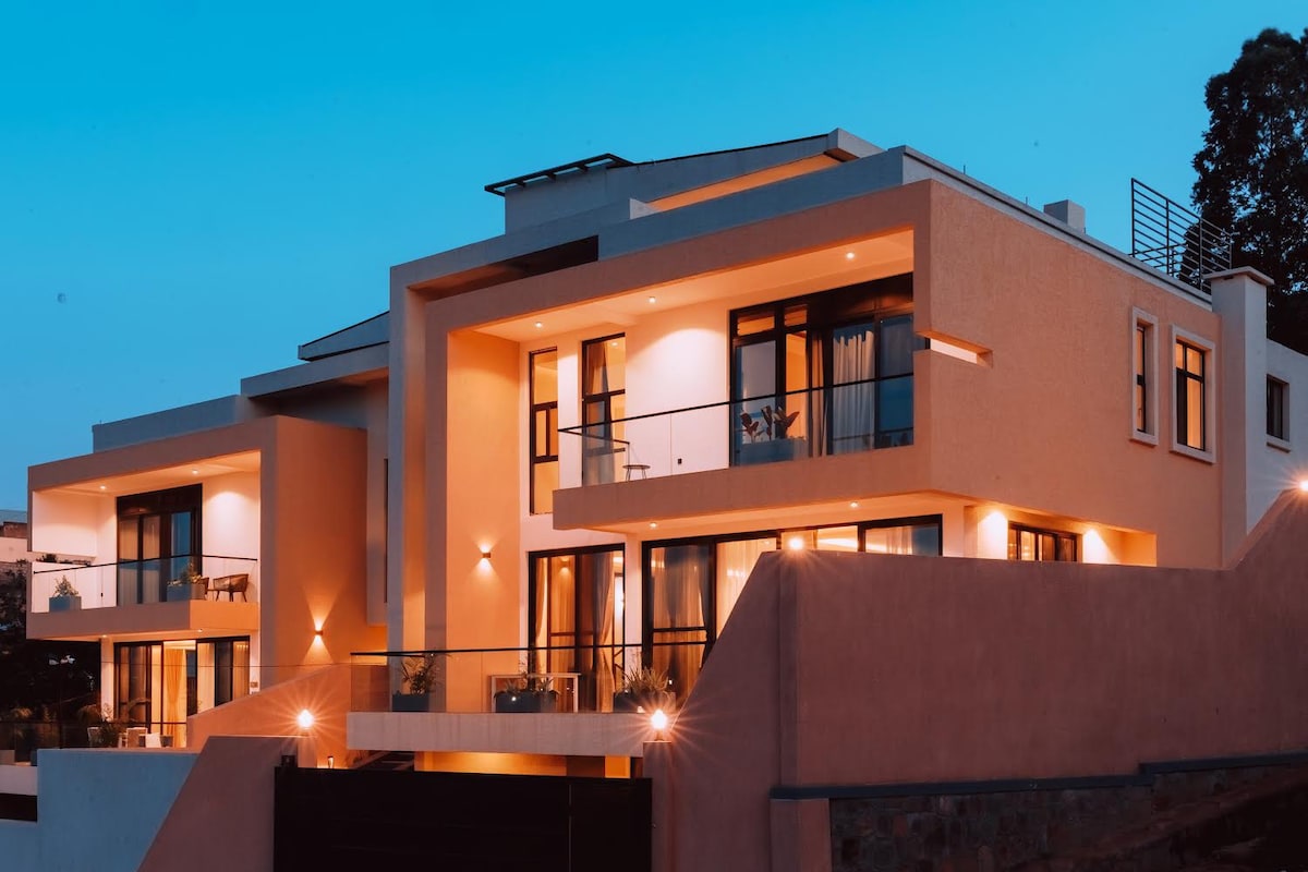The villa's exterior is showcased at dusk, featuring modern architecture with multiple recessed balconies and large windows. Soft outdoor lighting enhances the structure's design, while the rooftop terrace provides an elevated space for relaxation. The surrounding area is peaceful, contributing to a tranquil setting.