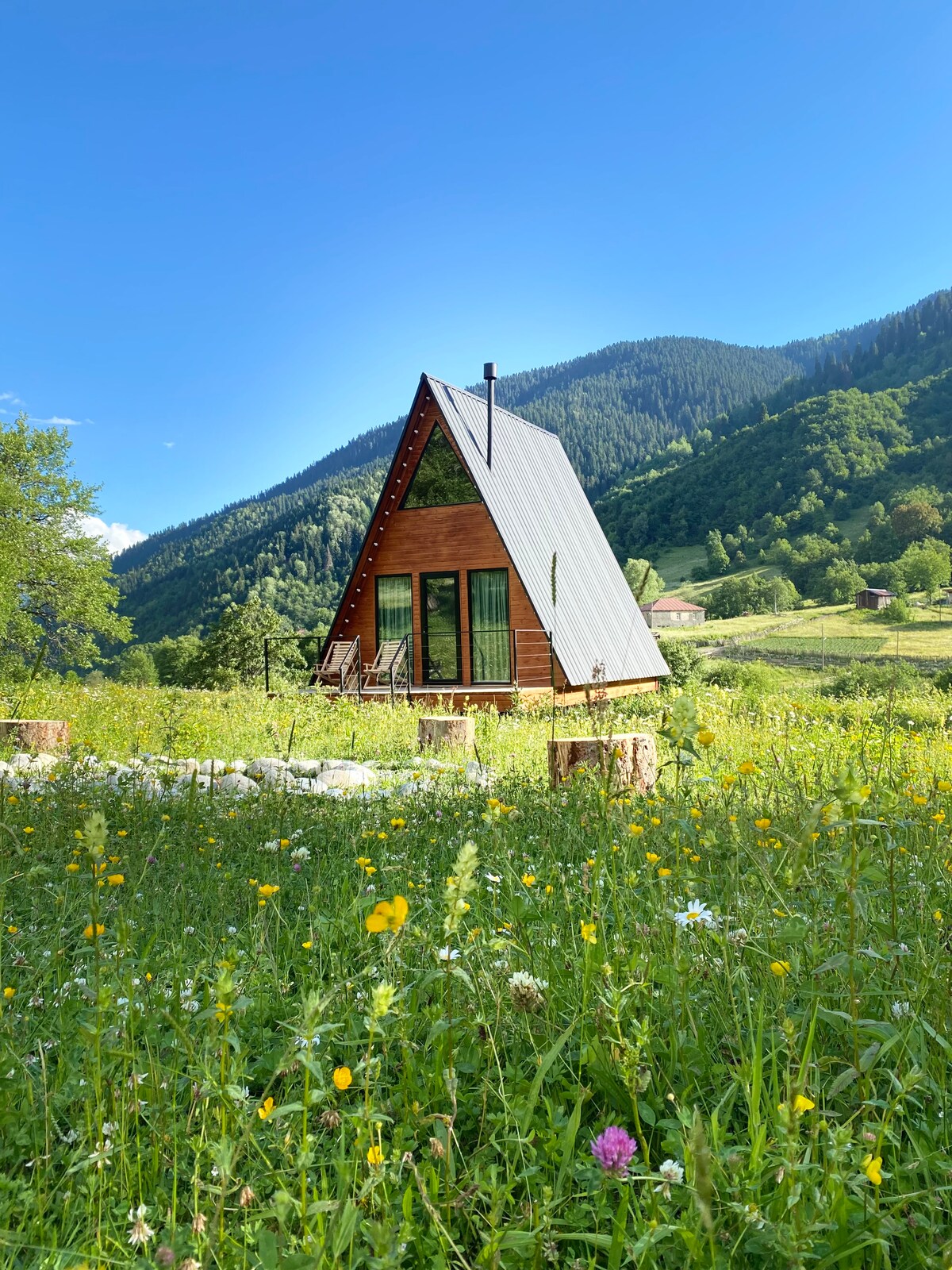 An A-frame cottage is positioned amidst a lush green field adorned with colorful wildflowers. The structure features large windows and a sloped roof, complemented by a forested backdrop of mountains. Sunlight illuminates the scene, creating a serene and inviting setting.