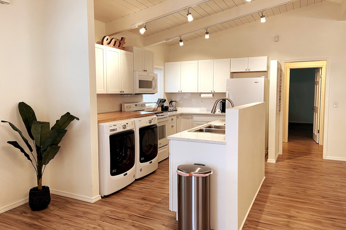 A modern kitchen is showcased with white cabinetry and a spacious countertop. Stainless steel appliances, including a washer and dryer, are visible. A potted plant adds a touch of greenery, while track lighting brightens the space, highlighted by light-colored walls and wooden flooring.