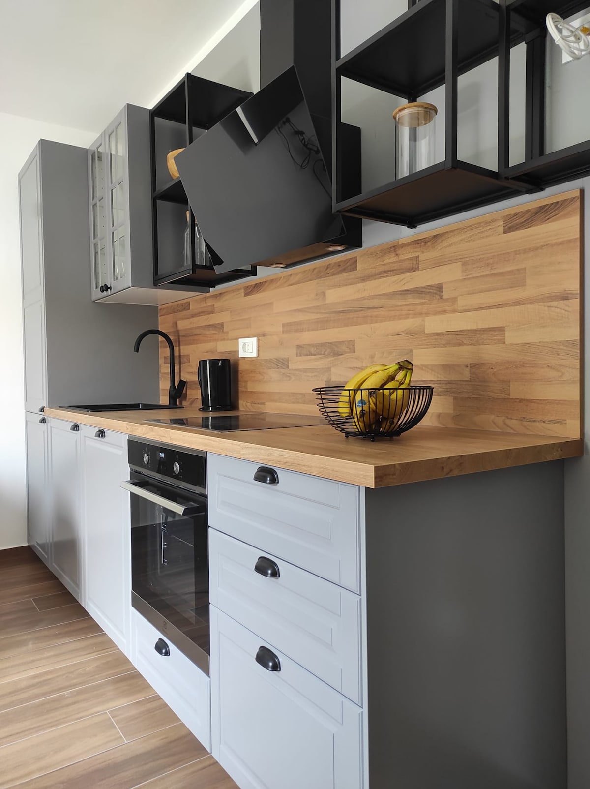 A modern kitchen is displayed with sleek cabinetry and a wooden countertop. A black coffee maker sits next to a decorative bowl containing bananas. An overhead cabinet showcases glassware, while a black faucet complements the minimalist design.