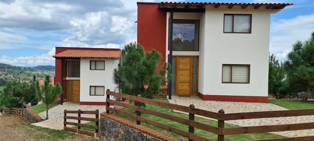 Two modern buildings feature a mix of red and white exteriors, surrounded by greenery. A stone pathway leads to the entrance, where wooden doors provide access. A wooden fence frames the property, complementing the natural landscape.