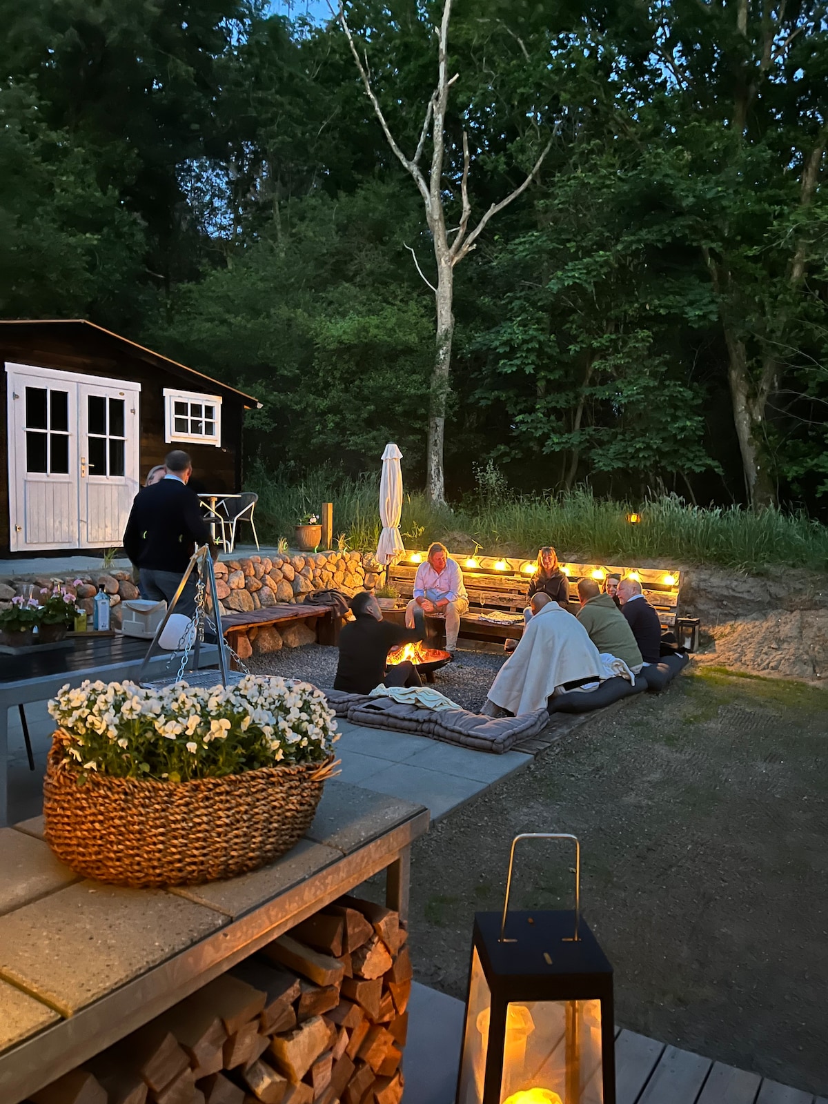 An evening setting showcases a cozy gathering around a fire pit, surrounded by seating with warm lighting. A rustic structure is visible in the background, framed by trees. Comfortable blankets and cushions create an inviting space, complemented by a flower basket and lantern nearby.