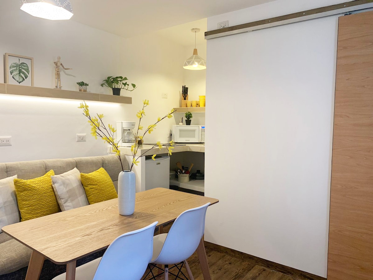 A light and airy dining area features a wooden table adorned with a vase of yellow flowers. In the background, a well-equipped kitchenette is visible, highlighted by modern appliances. Soft lighting is provided by a pendant lamp, enhancing the cozy atmosphere.