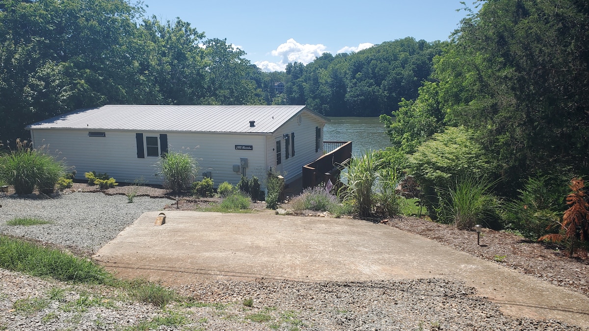 A serene lakefront home is surrounded by lush greenery and features a private path leading to the water. A gravel area in the foreground provides ample parking space, while the house is positioned slightly elevated, offering pleasant views of the lake and trees.