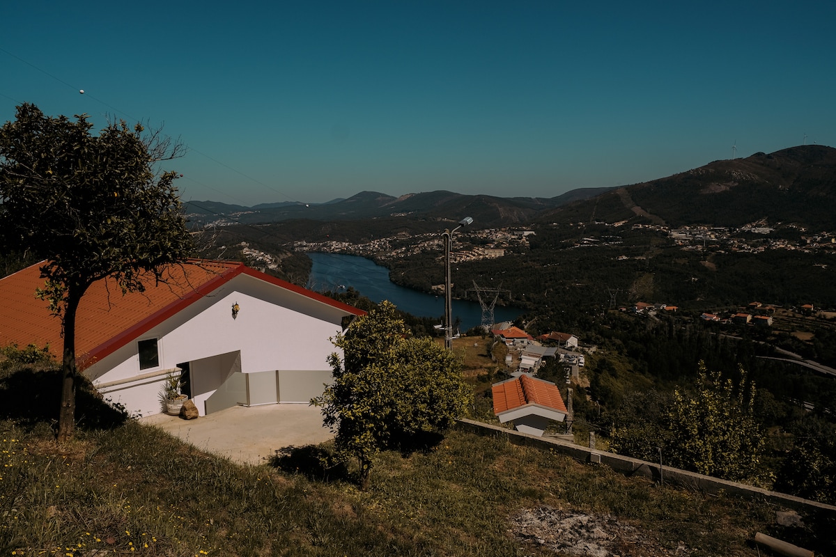 The exterior of the house is visible, featuring a white facade and red roof, set against a backdrop of rolling hills and the Douro River. Lush greenery surrounds the property, and the landscape extends in the distance, creating a serene rural atmosphere.