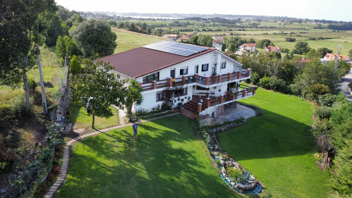 A two-story rural house is set on a spacious green lawn, surrounded by neatly arranged flower beds and pathways. Solar panels are visible on the roof, while balconies offer views of the surrounding landscape, which includes rolling fields and distant hills.