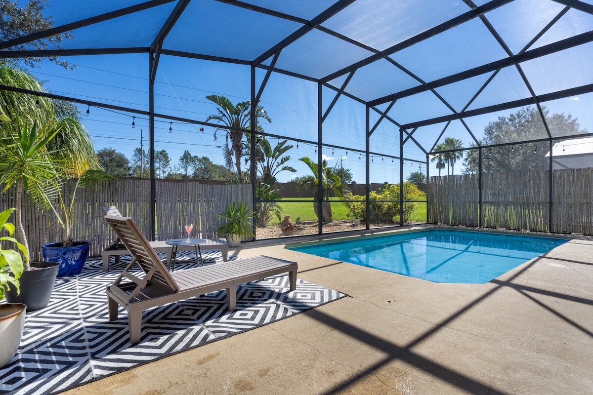 An outdoor pool area is enclosed by a mesh screen, featuring a heated swimming pool and lounge chairs. A geometric-patterned rug lies beside the pool, with pots of greenery adding a natural touch. Sunlight filters through, creating a bright and inviting space.