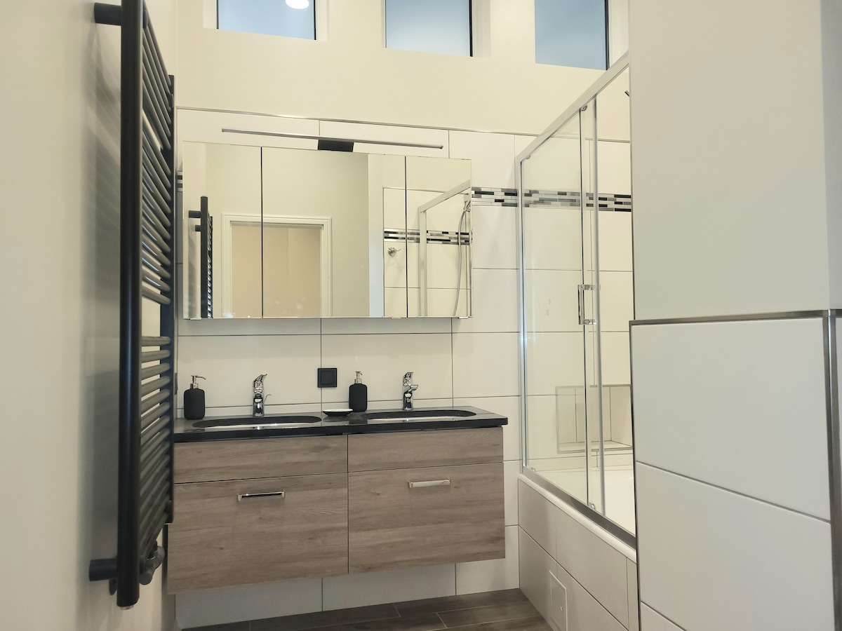 A modern bathroom features a sleek double vanity with a dark countertop and two sinks. A walk-in glass shower is visible alongside a bathtub. Natural light is provided by tall windows, while a stylish towel radiator is mounted on the wall.