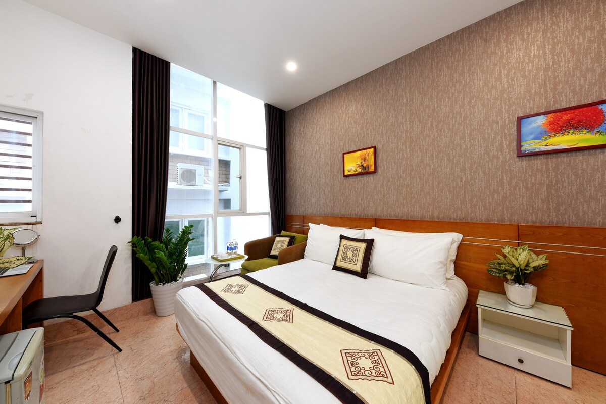 A well-appointed bedroom features a spacious bed with crisp white linens and decorative pillows. A small desk and chair are positioned beside a window, allowing natural light to brighten the space. Potted plants and wall art add a touch of greenery and color to the inviting atmosphere.