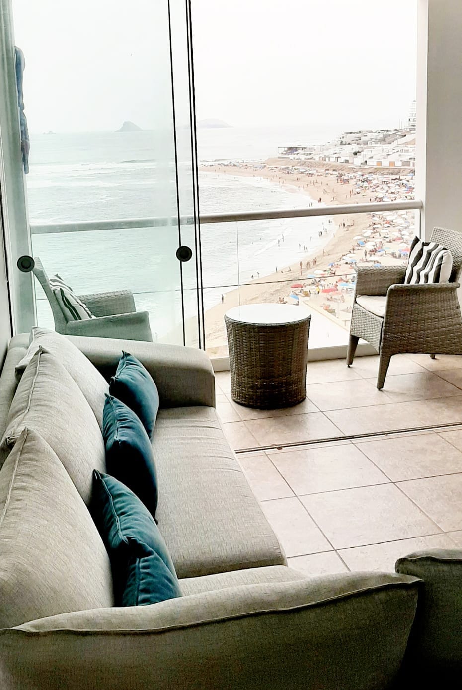 A modern seating area features a gray sofa with teal accent pillows, offering a view through large glass windows of the ocean and beach below. Two wicker chairs and a round side table complement the space, enhancing the relaxing atmosphere.