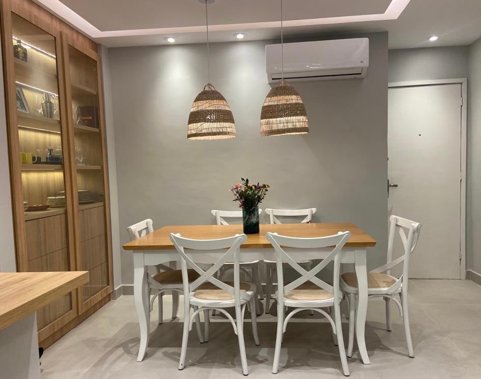 A dining area features a wooden table surrounded by six white chairs. Two woven pendant lights hang overhead, and a vase with flowers is placed in the center. The walls are painted in a neutral tone, creating a clean, modern atmosphere.