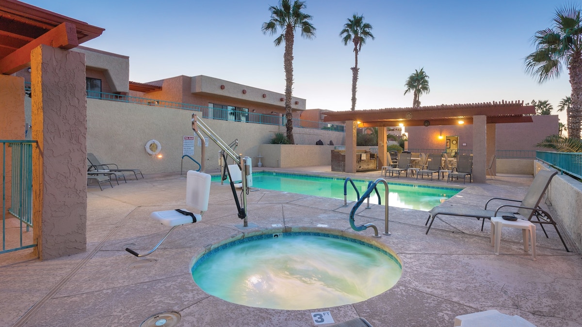 A swimming pool area features a clear blue pool alongside a relaxing hot tub. Palm trees provide shade, and a pergola offers covered seating. Several lounge chairs are arranged around the pool, and a lift for accessibility is positioned near the hot tub.