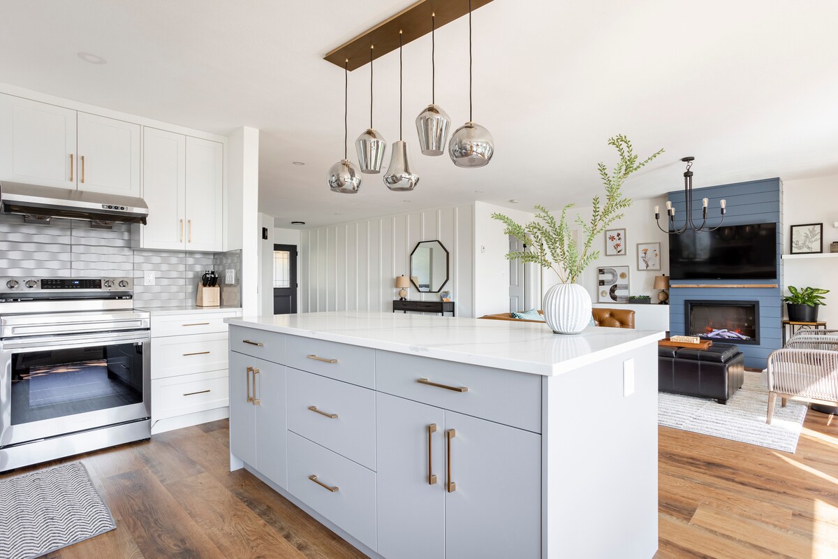 The kitchen features a large central island with sleek cabinetry and modern fixtures. Pendant lights hang above the island, illuminating the space. A well-equipped range and open living area are visible, with a stylish fireplace and comfortable seating creating an inviting atmosphere.