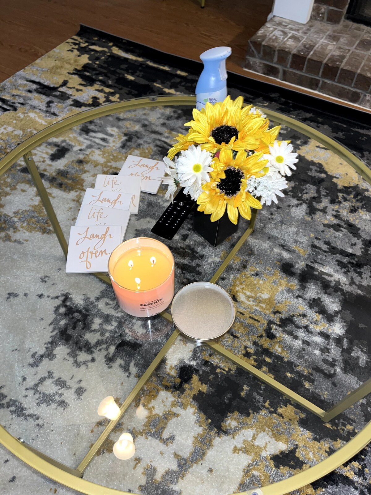 A glass coffee table is centered on a patterned rug, accented by a decorative arrangement of sunflowers and daisies in a vase. A lit candle and a scented spray are placed on the table, alongside neatly arranged note cards with uplifting phrases.