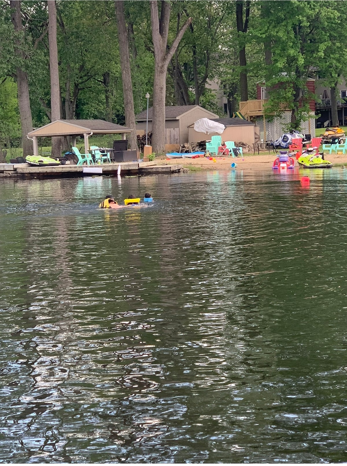 A calm lake scene is depicted, with floating objects in the water. A gazebo is visible on the shore, accompanied by colorful lounge chairs and various inflatable toys positioned on the sandy beach. Tall trees frame the background, creating a serene atmosphere.