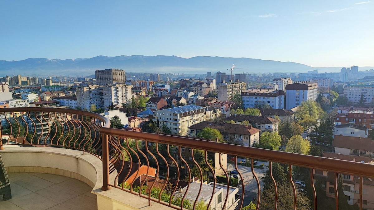 A panoramic view showcases a vibrant cityscape, featuring a mix of residential and commercial buildings, with lush greenery interspersed. The distant mountain range frames the horizon under a clear blue sky, while a railing adds a structural element to the foreground.