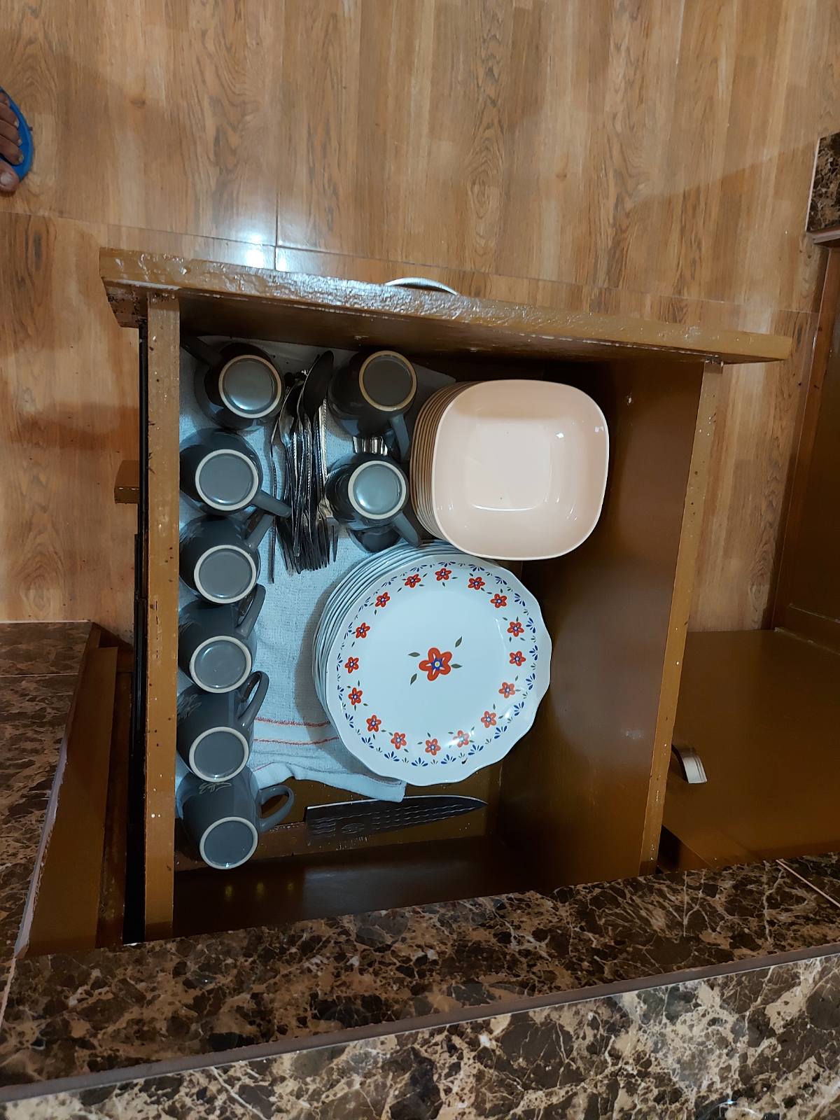 A kitchen drawer is displayed, containing an organized assortment of dishware. The collection includes a stack of plates adorned with a floral design, a bowl, cups, and various utensils. A white towel is positioned below the items for convenience.
