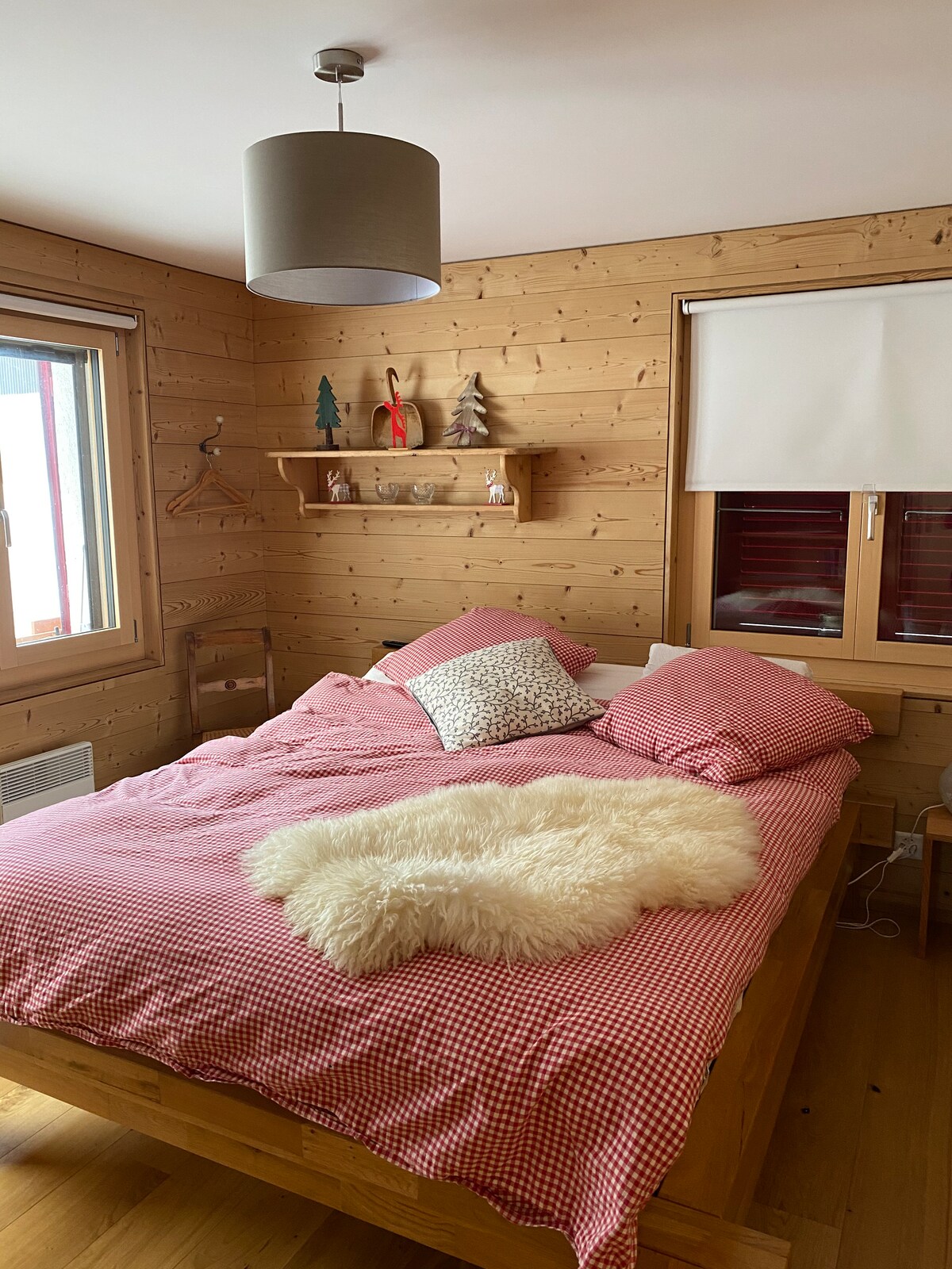 A cozy bedroom features a spacious bed with red checkered bedding and decorative pillows. Soft texture is added by a plush white throw draped over the bed. Wooden walls create a warm atmosphere, complemented by natural light filtering through the window with a light shade.