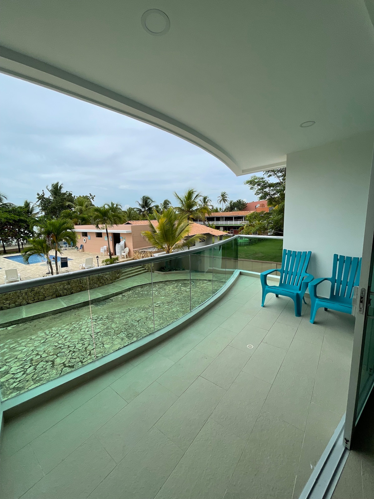 A private balcony features two bright blue chairs overlooking lush greenery and a pool area. The modern glass railing enhances the open feel, allowing for unobstructed views of the surroundings, including palm trees and nearby buildings.