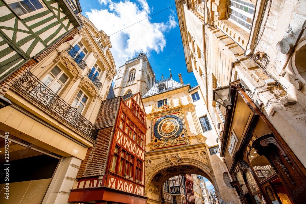 An upward view of a narrow street in Rouen features historic half-timbered buildings and a large, ornate clock. The bright blue sky peeks through the clouds, illuminating the intricate architecture and details of the structures lining the street, creating a charming ambiance.