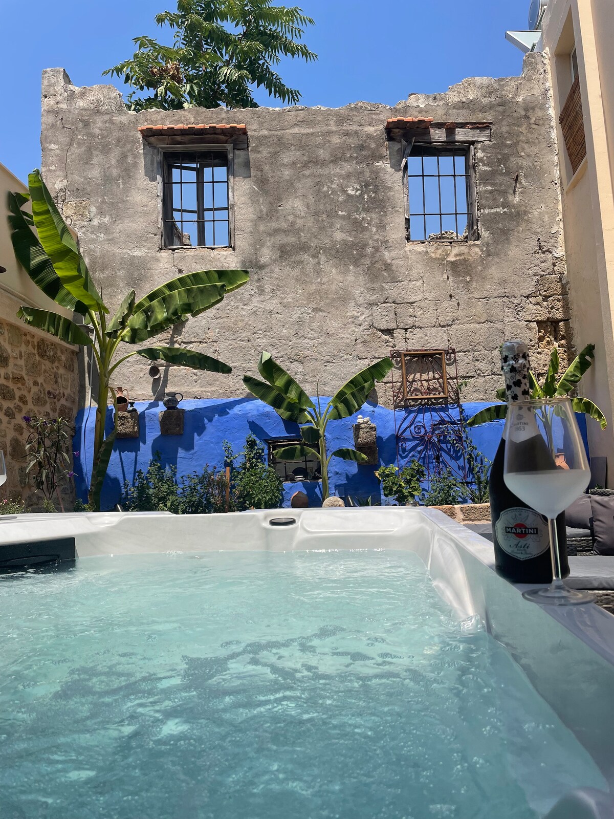 A hot tub is visible in the foreground, its water reflecting sunlight. Behind, a weathered stone wall features broken windows and vibrant blue accents. Tropical plants and greenery surround the area, adding a touch of nature to the inviting outdoor space.