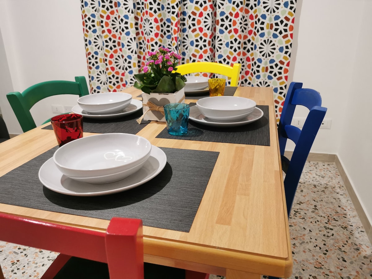 A dining table is set with white plates and glassware in vibrant colors, surrounded by multi-colored chairs. A decorative potted plant adds a touch of greenery. Colorful patterned curtains provide a cheerful backdrop, enhancing the inviting atmosphere of the dining space.