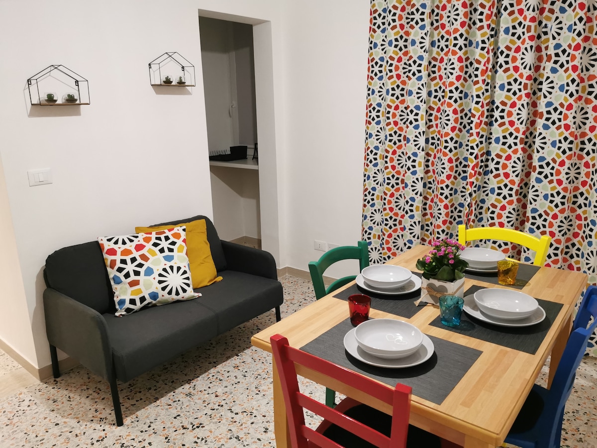 A dining area is featured with a wooden table set for four, accompanied by colorful chairs in red, yellow, and blue. A cozy gray sofa with decorative cushions is positioned nearby. The window is adorned with vibrant patterned curtains, enhancing the lively atmosphere of the space.