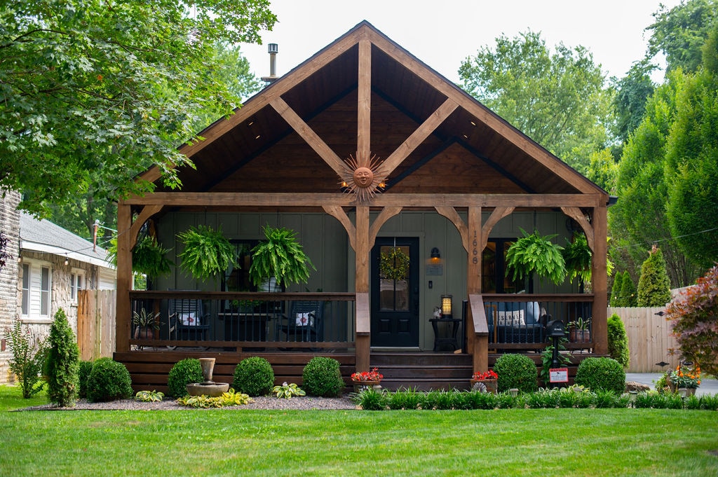 A charming exterior of a single-story home is shown, featuring a covered porch supported by wooden beams. Lush ferns hang from the railing, and landscaped greenery enhances the welcoming appearance. A well-manicured lawn leads to the entrance, inviting relaxation and enjoyment of the surrounding nature.