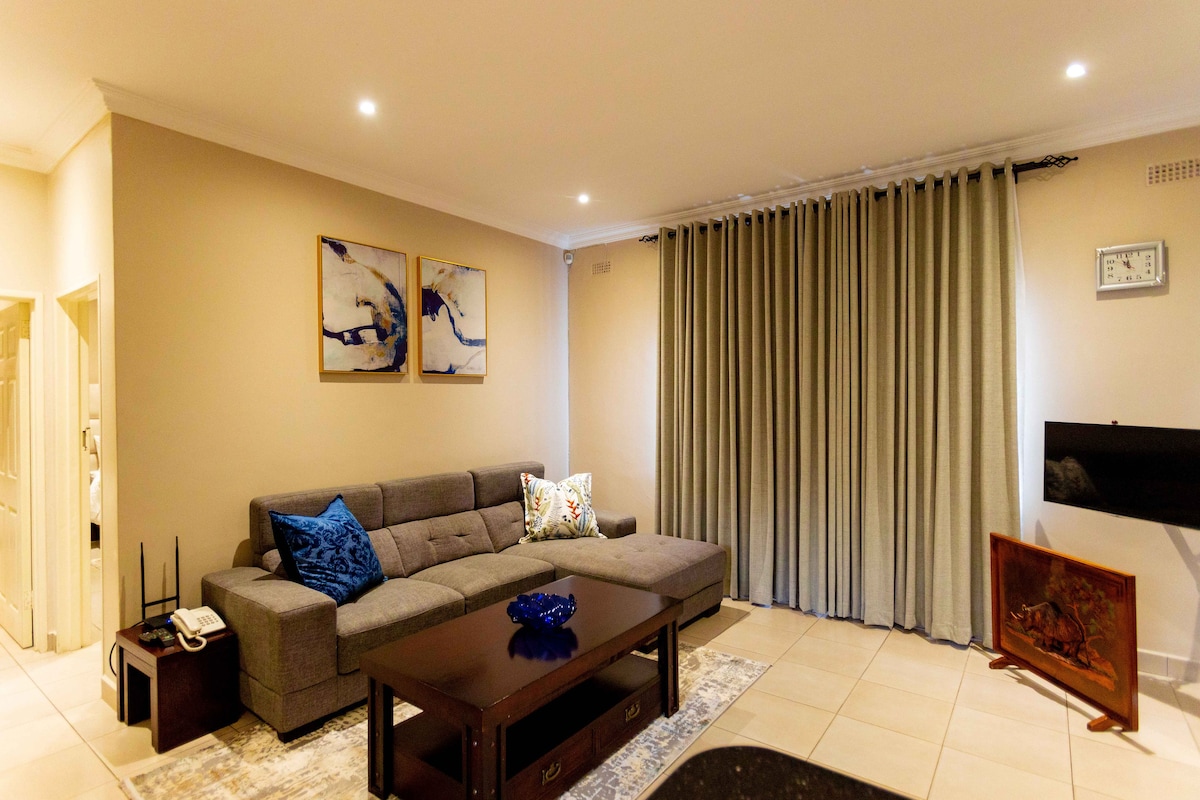 A modern living area features a comfortable grey sectional sofa adorned with colorful throw pillows. Light-colored walls are complemented by two framed artworks. Floor-to-ceiling curtains provide privacy, while subtle ambient lighting adds to the inviting atmosphere. A sleek coffee table sits below a decorative centerpiece.