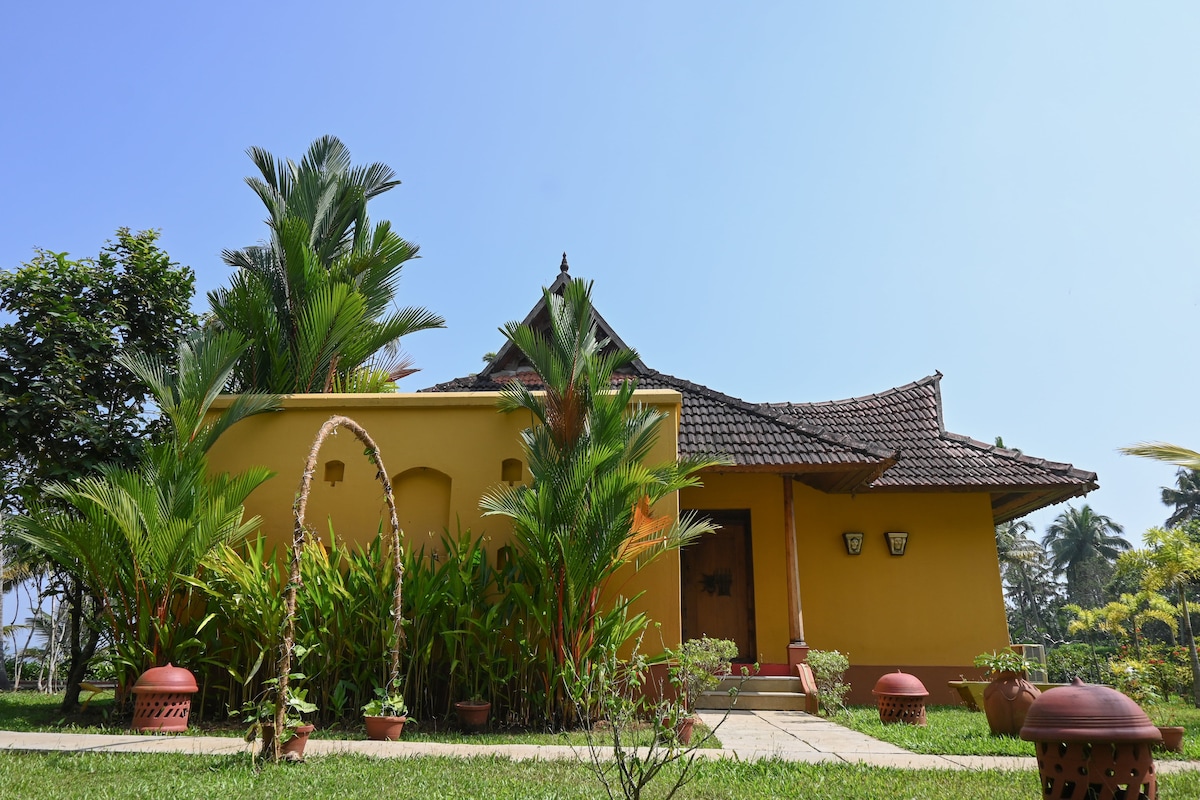 A vibrant yellow house is surrounded by lush greenery, including tall palms and ornamental plants. The entrance features a paved path leading to the doorway, adorned with two wall-mounted lights. Colorful pots line the garden, contributing to the welcoming ambiance of the property.