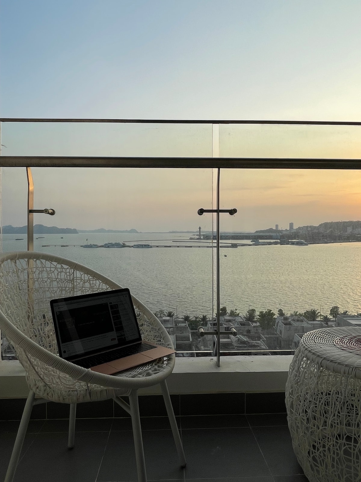 A balcony scene is presented with a laptop positioned on a round table. The view includes a serene waterfront illuminated by the warm hues of sunset. Curved white chairs provide a comfortable setting for relaxation while overlooking the scenic landscape.