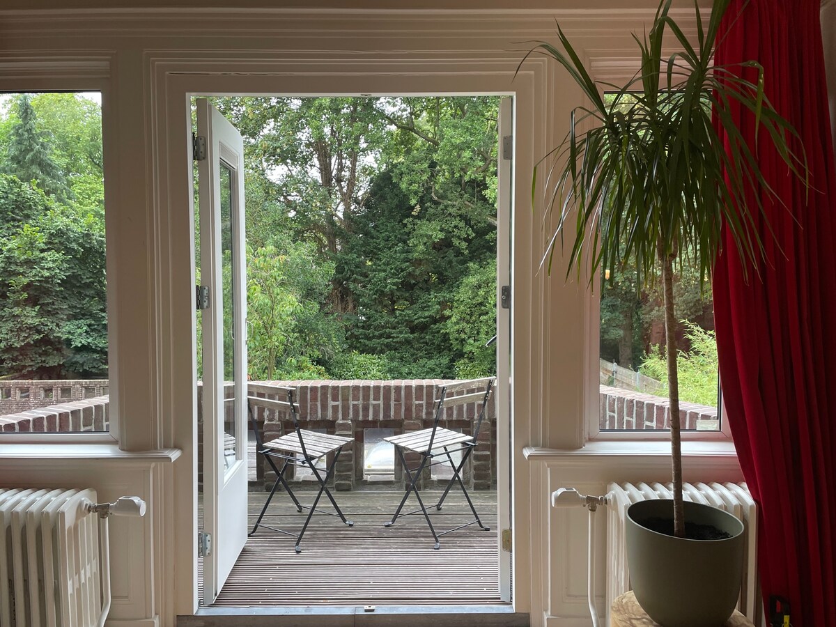 A view from inside the villa shows double doors opening to a private terrace surrounded by lush greenery. Two folding chairs are positioned on the wooden deck, creating an inviting outdoor seating area framed by tall plant life and a bright red curtain.
