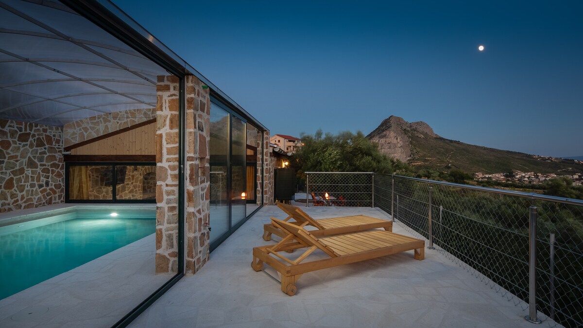 An indoor pool with clear blue water is visible, surrounded by a natural stone wall. Two wooden sun loungers are positioned on the terrace. A mountain can be seen in the background, and a full moon casts a gentle glow over the scene.