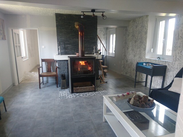 A welcoming living area features a decorative fireplace with a Modern flue and is surrounded by light-colored walls with patterned accents. Two windows allow natural light, and seating options include a dark chair and a black armchair. A coffee table with decorative elements sits at the center.