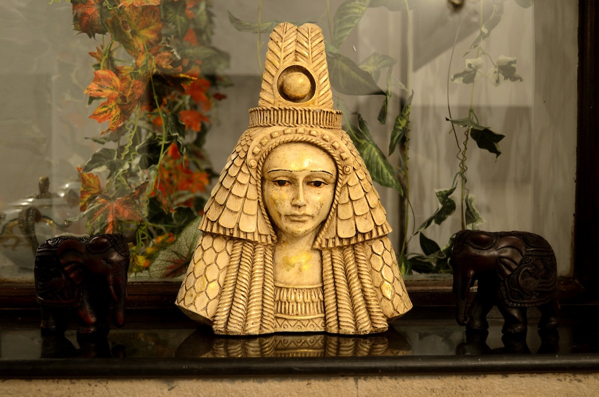 A decorative sculpture is prominently displayed against a glass backdrop. The sculpture exhibits intricate detailing with a textured surface. Two small wooden elephant figurines are positioned on either side, complementing the decorative piece, while greenery is softly visible in the background.