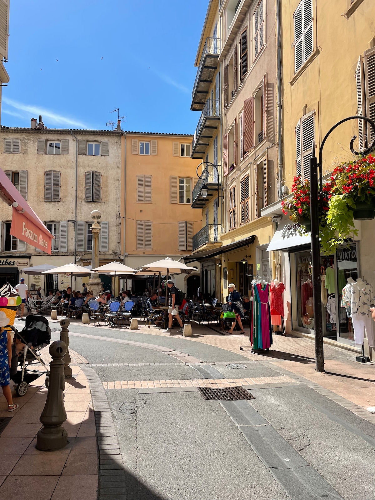 A bustling street scene captures the charm of old Antibes. Cafe terraces are filled with patrons enjoying the sunny weather. Shop displays line the sidewalk, showcasing colorful clothing and vibrant flower arrangements hang from street lamps, adding to the lively atmosphere.