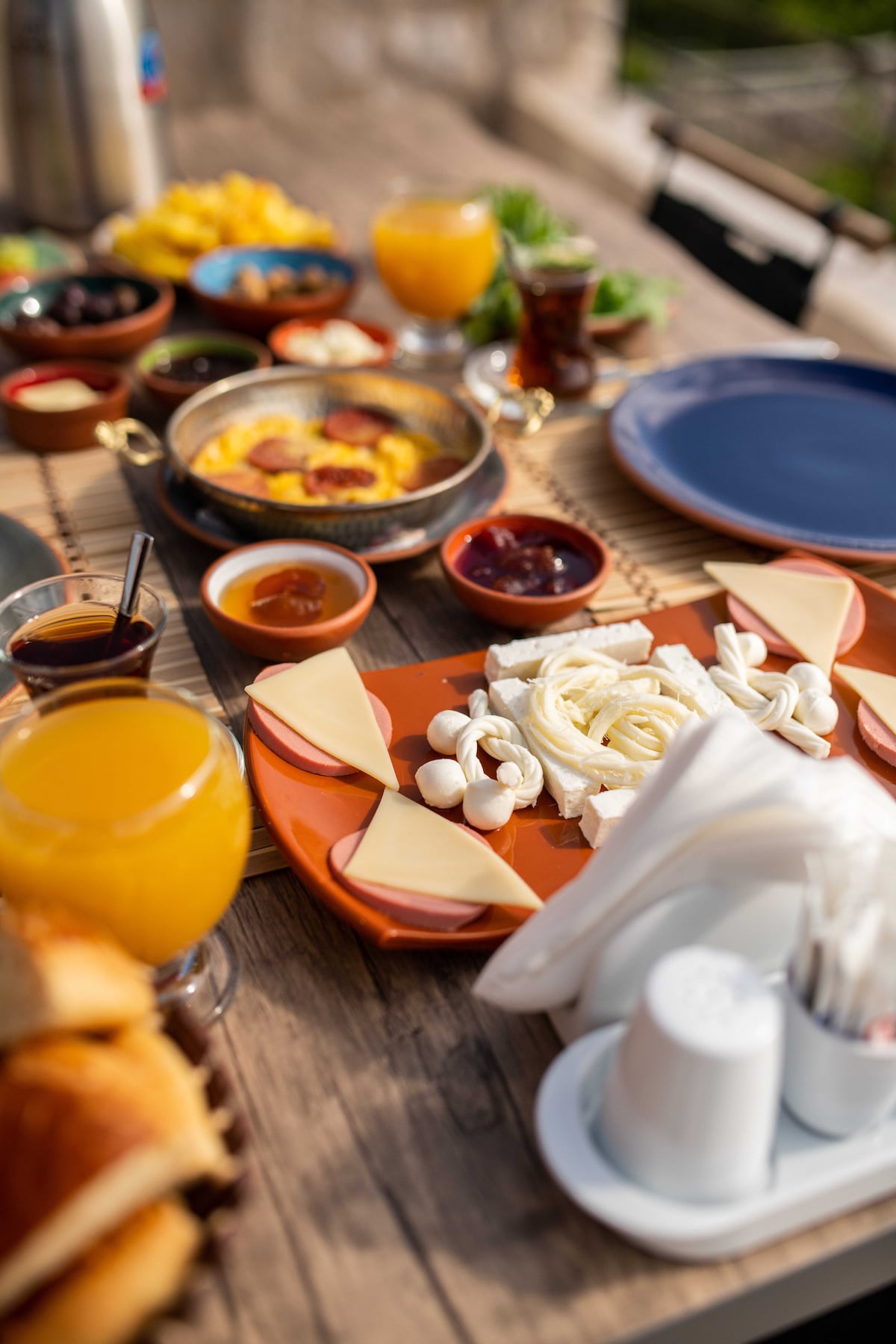 A wooden table is set for breakfast, featuring a variety of colorful dishes. Fresh cheeses and olives are artfully arranged on an orange plate, accompanied by bowls of jams and honey. Two glasses of orange juice and hot tea complete the inviting spread.