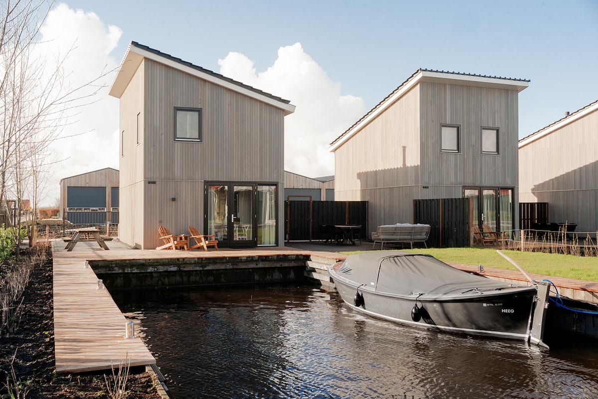 Two modern waterlodges are depicted, featuring wooden exteriors and large glass doors. A wooden dock extends towards a moored boat, with seating arrangements visible on the terrace. The surrounding landscape is open, enhancing the connection to the water.