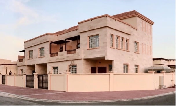 A two-story villa is depicted with a beige stone exterior and multiple balconies. Large windows are featured on both levels, allowing ample light. A gated entrance is visible, and the surrounding area is paved, suggesting easy access. The structure is set against a clear blue sky.