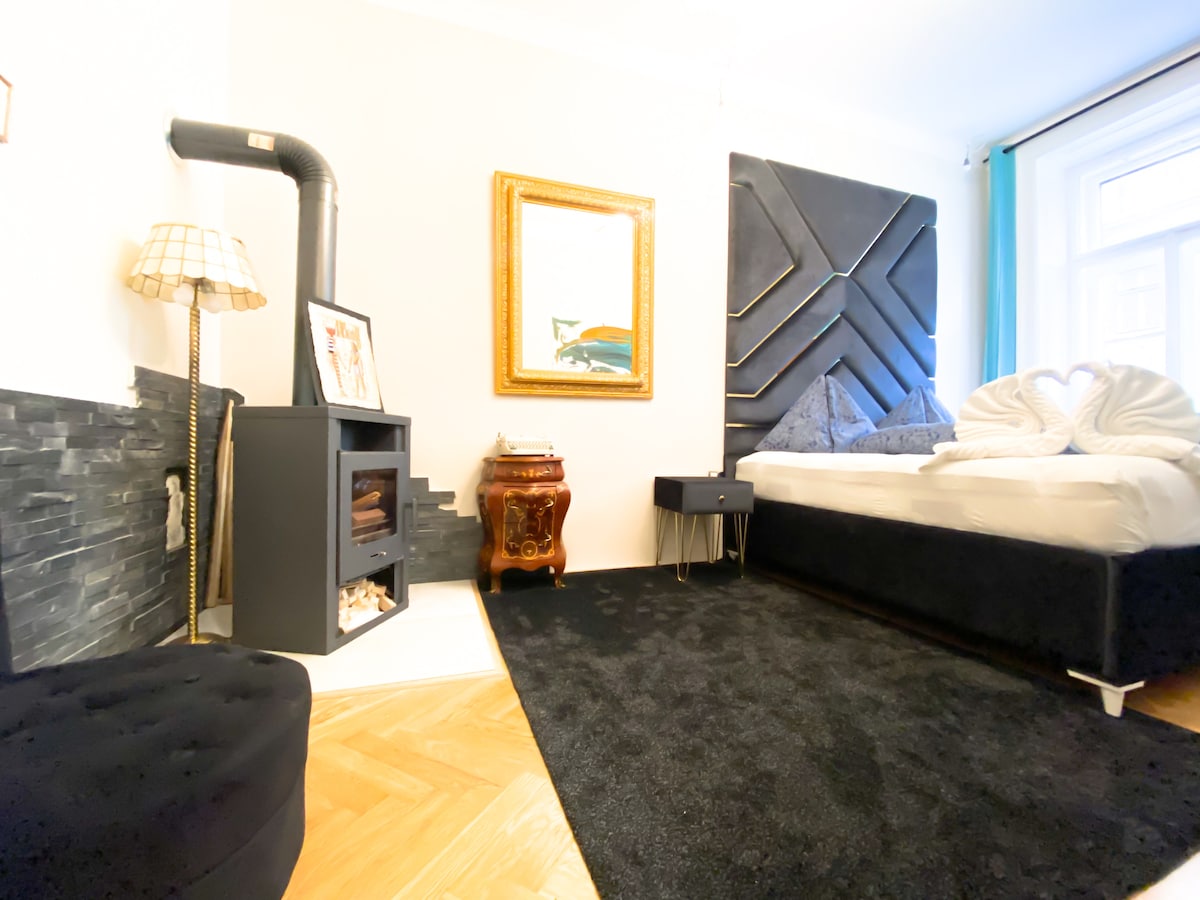 A cozy sleeping area is shown, featuring a spacious bed with decorative pillows. A black and gray accent wall is present, alongside a stylish seating area. A vintage lamp and decorative frame complement the space, while a window allows natural light to enter.