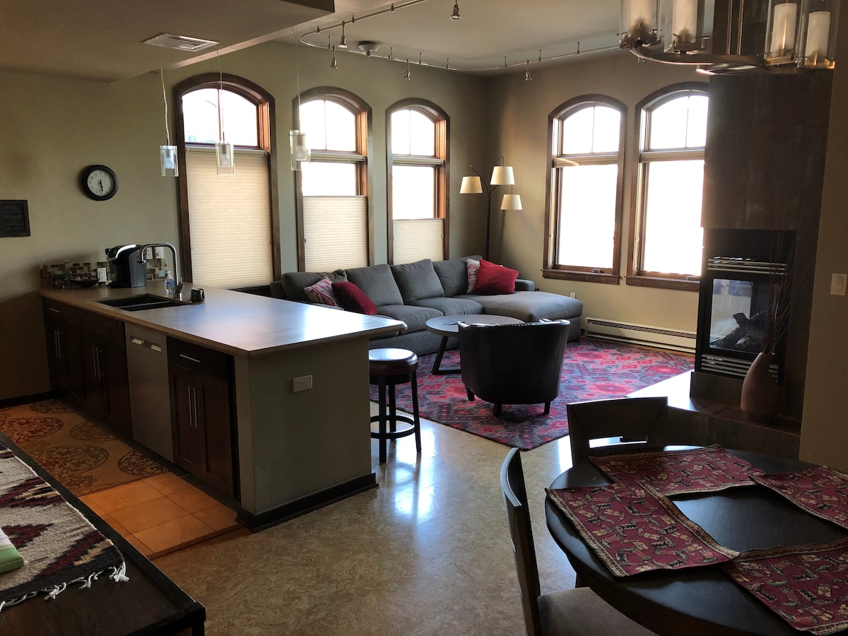 A spacious living area features a plush sectional couch adorned with pillows, positioned near large arched windows. A modern fireplace stands in the corner, complemented by a circular dining table and sleek kitchen counter. Decorative rugs add warmth to the open space, enhancing its inviting atmosphere.