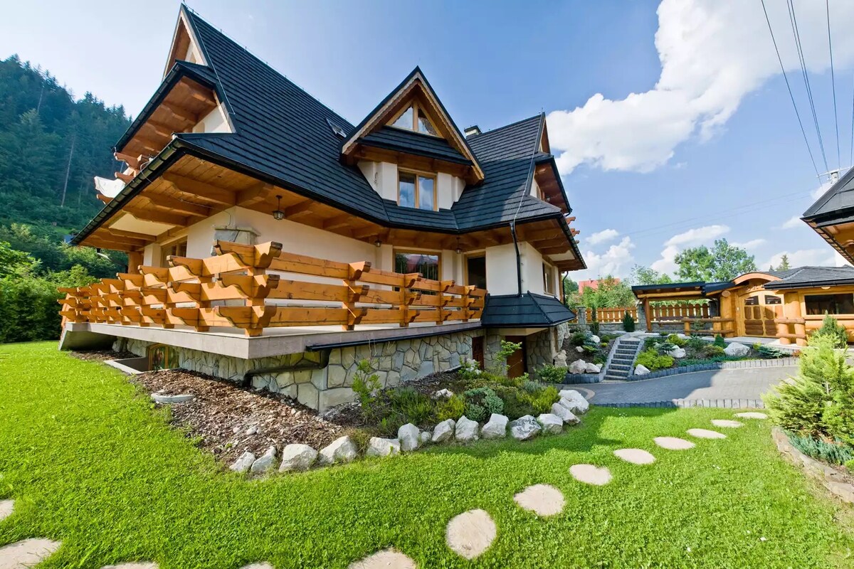 A charming wooden structure with a steeply pitched roof is situated amidst lush greenery. A spacious terrace with a wooden railing extends from the front, complemented by a well-maintained garden featuring stone pathways and decorative boulders leading to additional outdoor areas.
