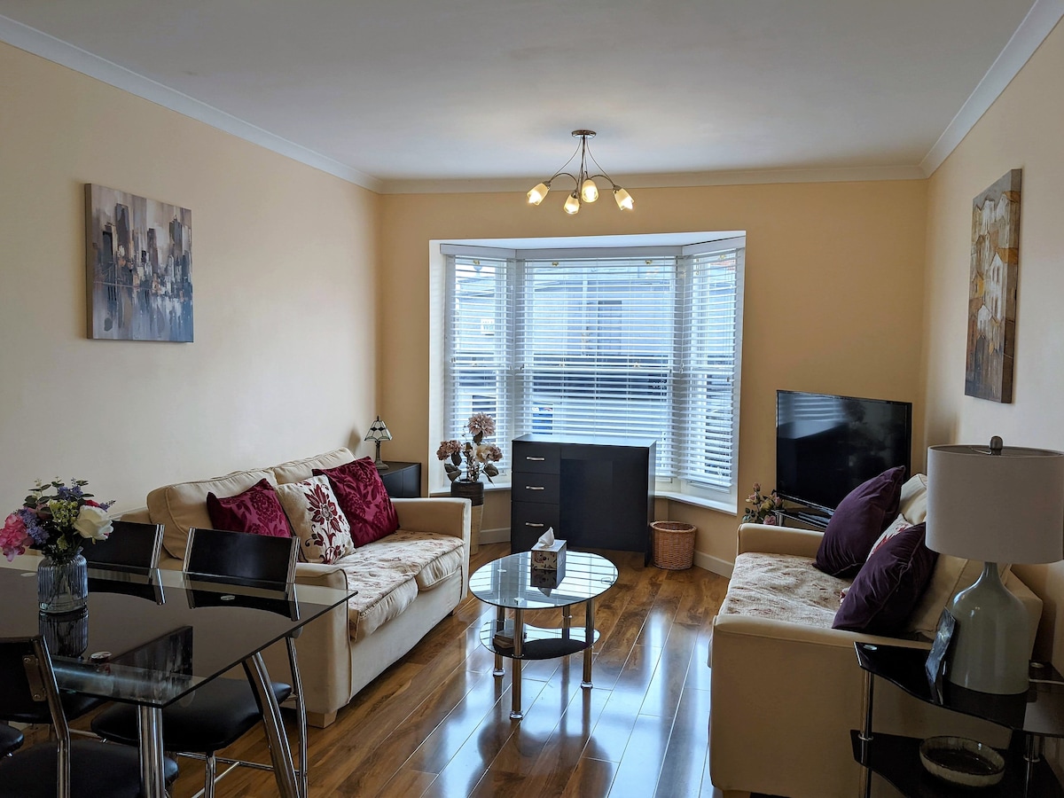A comfortable living area is presented with light walls and stylish decor. Two sofas are arranged around a glass coffee table, while a flat-screen TV sits against a wall. Natural light streams in through the bay window, enhancing the inviting space.