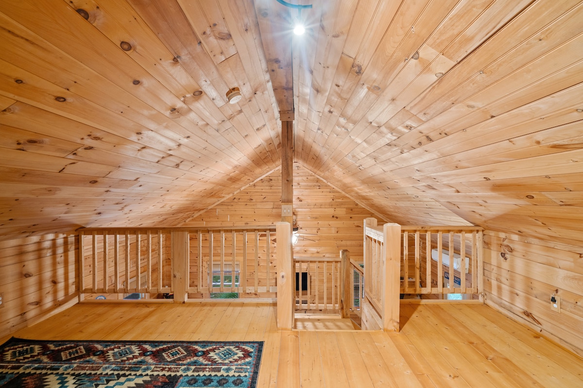 A wooden loft area features sloped ceilings and natural wood paneling throughout. The space includes a wooden railing overlooking the lower level, with a small, colorful area rug positioned on the floor. Soft lighting enhances the inviting feel of the loft.
