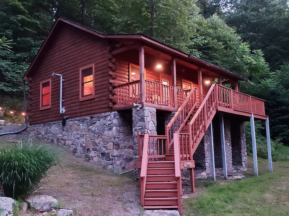 A rustic log cabin is set amidst lush greenery, featuring a stone and wooden exterior. Sturdy stairs lead up to a spacious front deck, where warm lights create a welcoming ambiance. The surrounding nature offers a serene backdrop for relaxation.