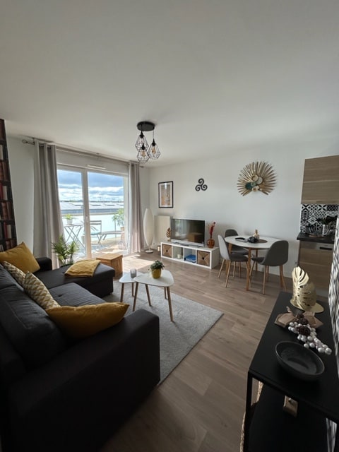 A bright living area features a comfortable sofa adorned with decorative pillows. A coffee table sits on a soft rug, while a modern TV unit displays a television. A dining table with stylish chairs is positioned next to large glass doors, providing access to a balcony with scenic views.