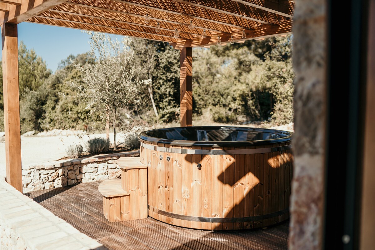 An outdoor hot tub made of wood is nestled under a wooden pergola. Steps lead up to the hot tub, which is bordered by natural stone walls. Surrounding greenery enhances the serene atmosphere, providing a secluded setting for relaxation.
